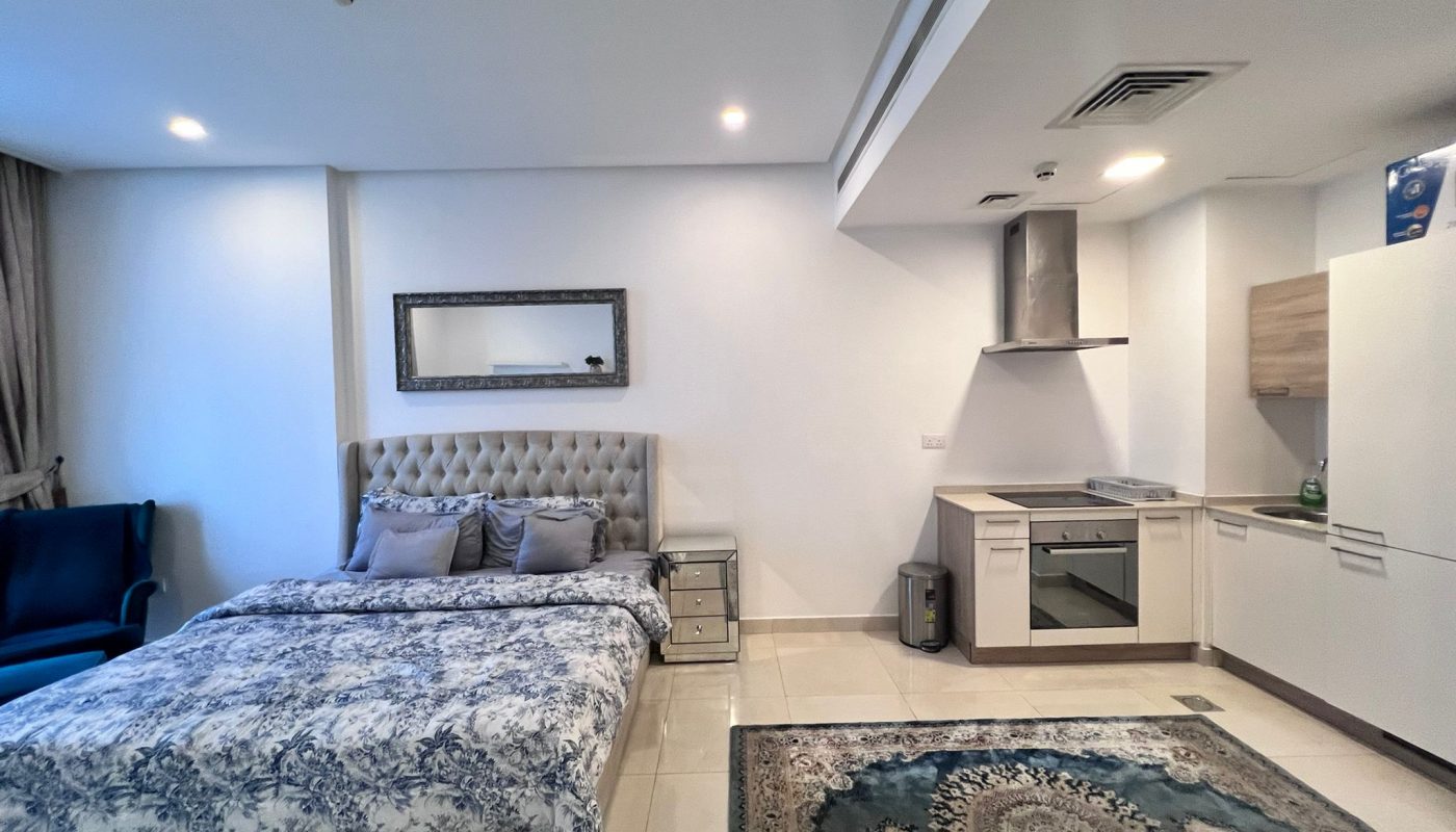 A modern studio apartment with a bed, nightstand, armchair, and a kitchenette. The room features a wall mirror and a patterned rug on a tiled floor.