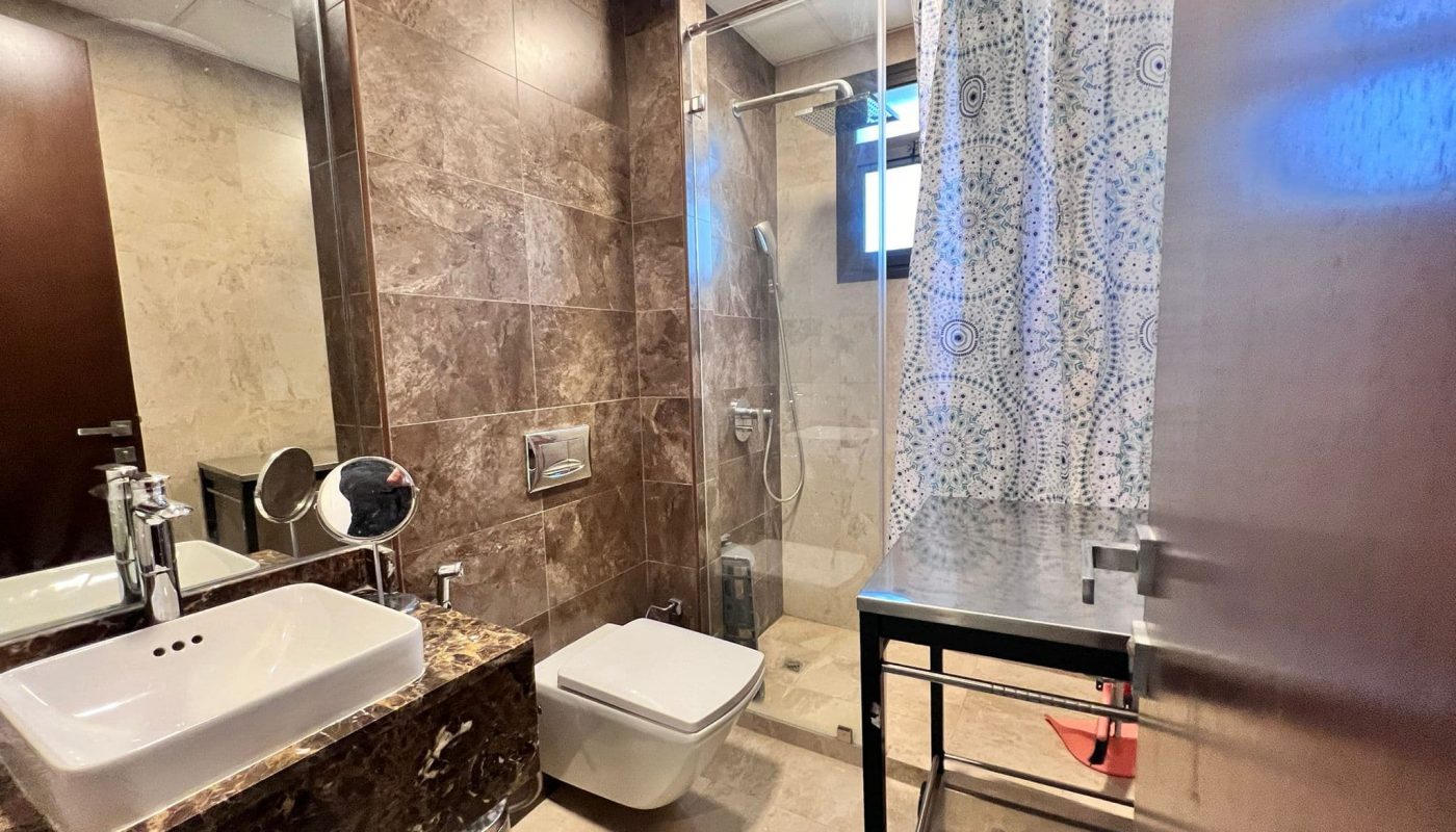 Modern bathroom with a sink on a marble countertop, wall-mounted toilet, walk-in shower, and patterned curtain. Brown tiled walls and a metal storage rack visible.