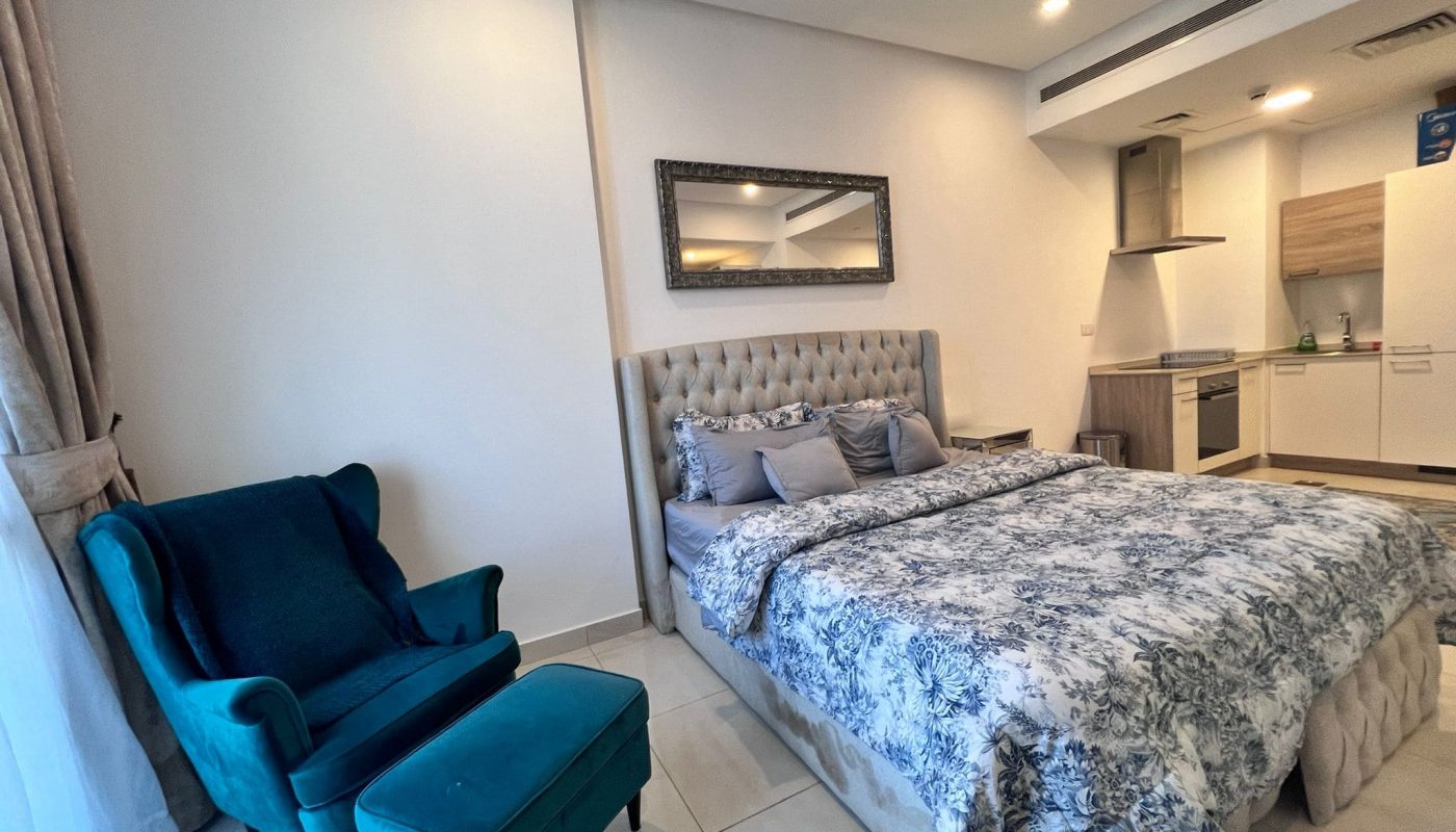 Bedroom with a tufted headboard bed, blue armchair, matching ottoman, and a mirror. A small kitchen area is visible in the background.