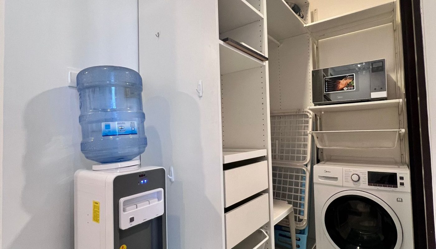 Laundry room with a white washer, open shelving, blue mat, water dispenser, and microwave on a shelf.