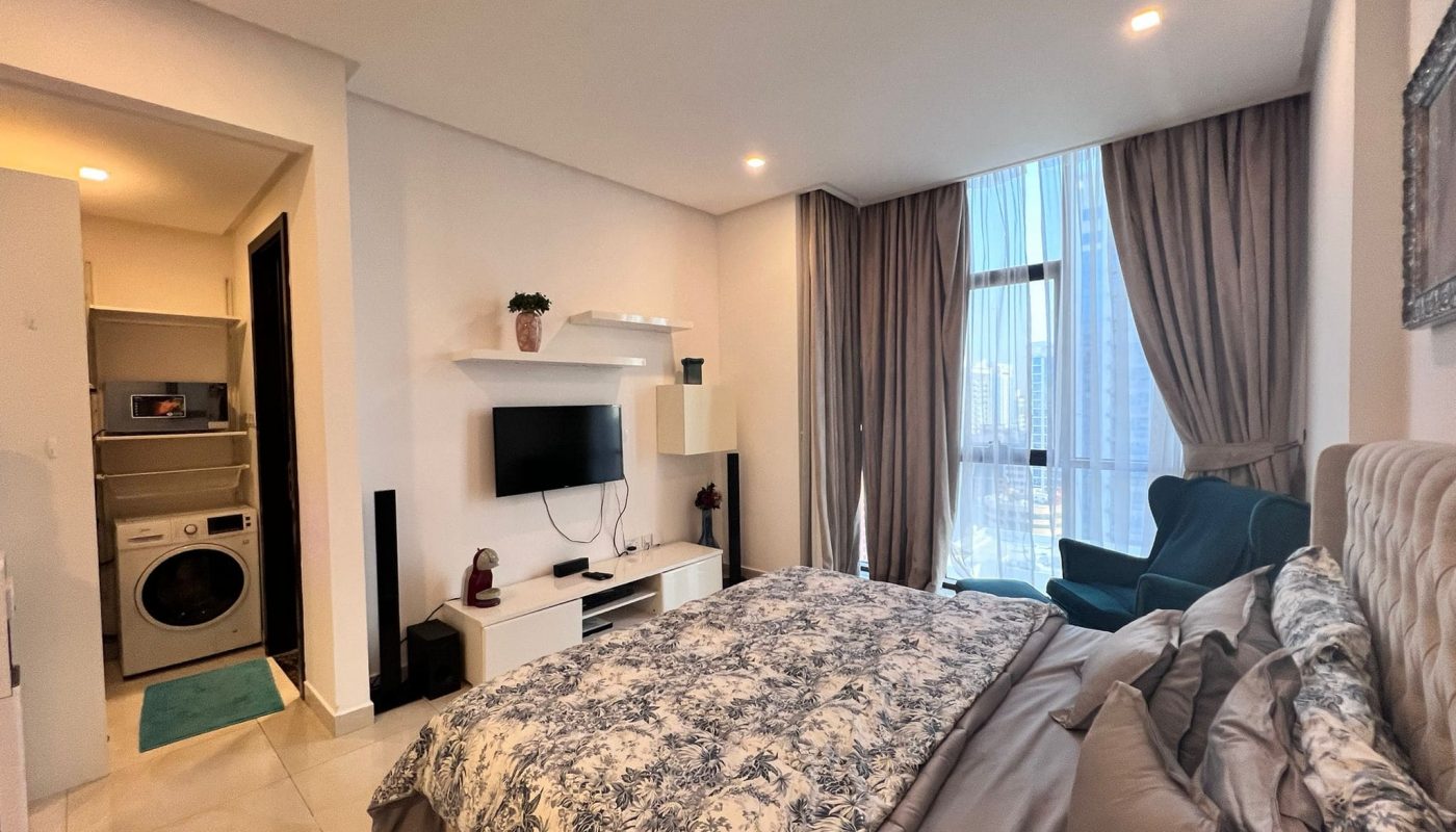 Modern bedroom with a floral bedspread, wall-mounted TV, armchair, and large window with curtains. A small laundry area with a washer and dryer is visible through an open door.