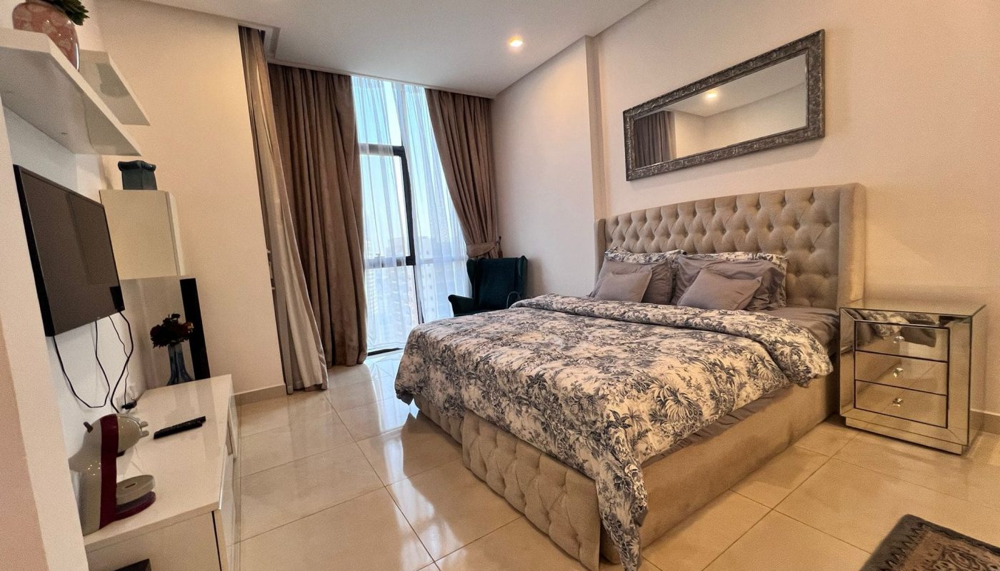 A modern bedroom with a king-size bed, patterned bedding, a wall-mounted TV, a mirrored nightstand, and floor-to-ceiling windows with curtains.