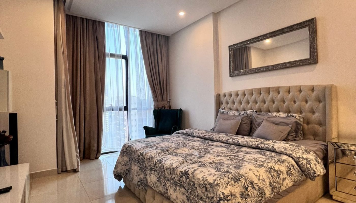 Modern bedroom with a large bed, patterned bedding, and upholstered headboard. Room features a wall mirror, floor-to-ceiling curtains, a chair, and neutral-colored tiles.