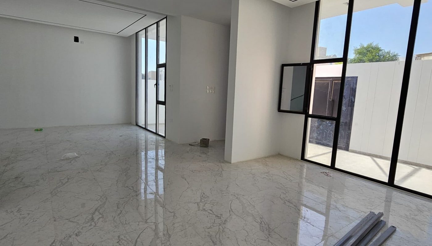 A spacious room with large glass windows, a marble floor, and a partially installed glass door. A cardboard box and construction materials are on the floor.