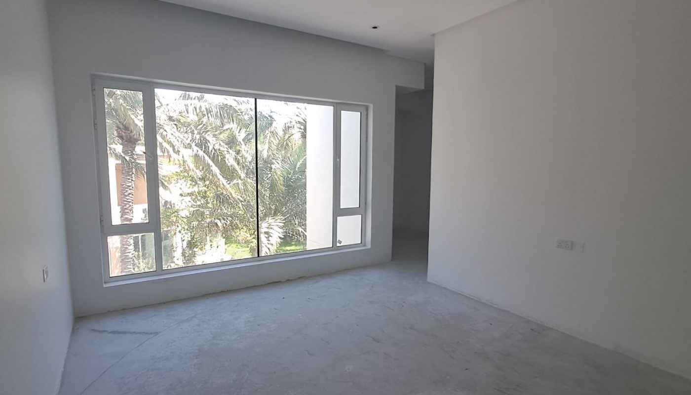 An empty, unfurnished room with white walls, a large window overlooking trees, and an unfinished concrete floor.