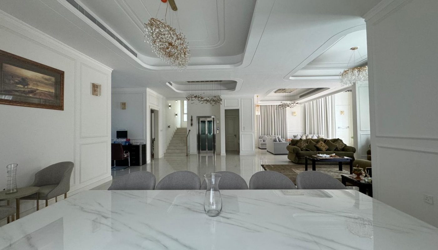 Spacious modern living area with a marble dining table, gray chairs, elegant chandeliers, a seating area with green sofas, and a staircase leading upstairs.