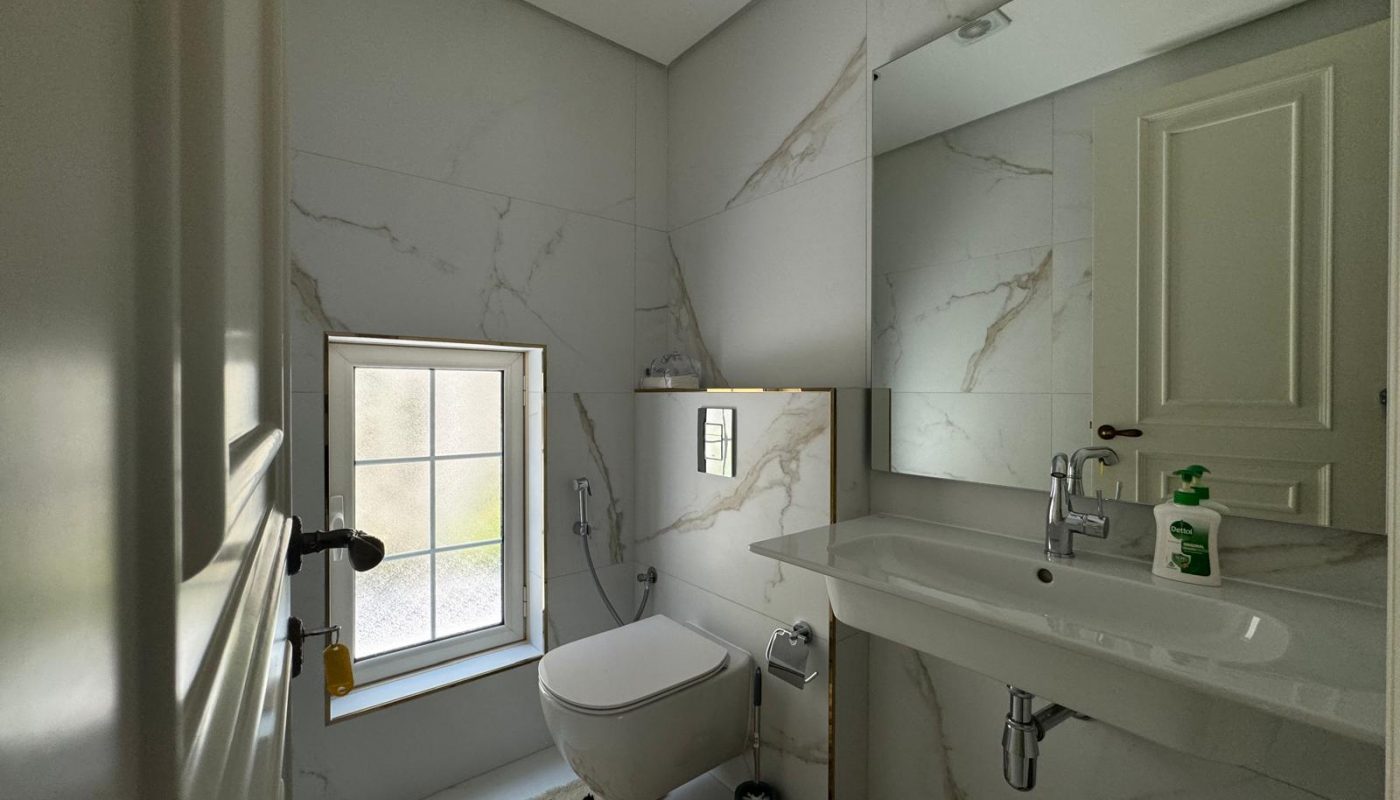 Modern bathroom with white marble walls, a wall-mounted toilet, sink with mirror, and a small window. Soap dispensers are on the sink.