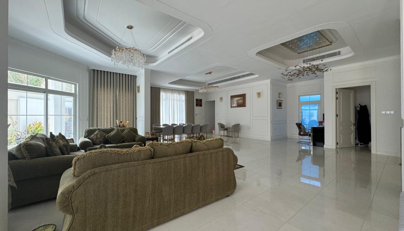 Spacious living room with green sofas, tile flooring, and a large dining table. Chandeliers hang from a coffered ceiling. Large windows allow natural light. Art decorates the white walls.