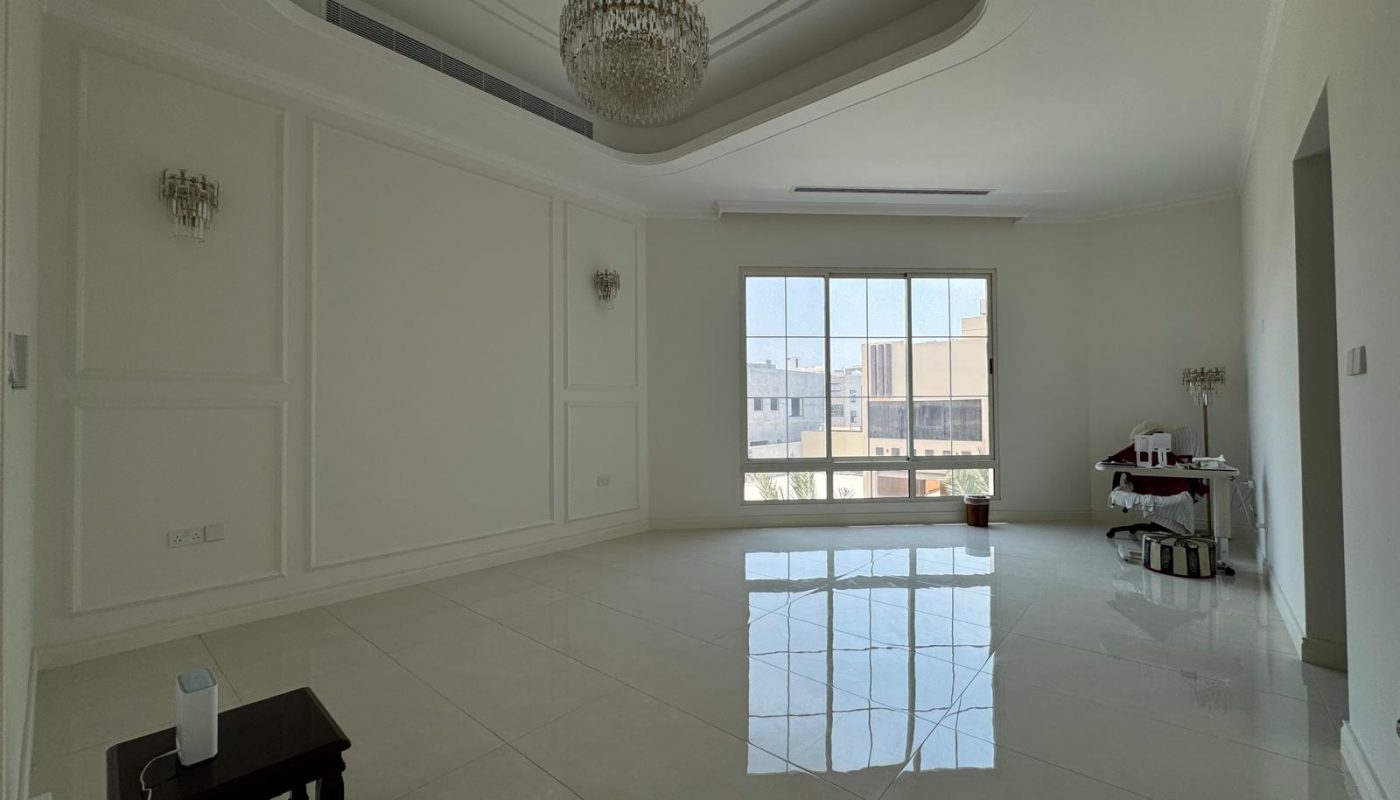 An empty, unfurnished room with white walls, glossy tiled floor, chandelier, and large window. A small table with items and bags is in the corner.