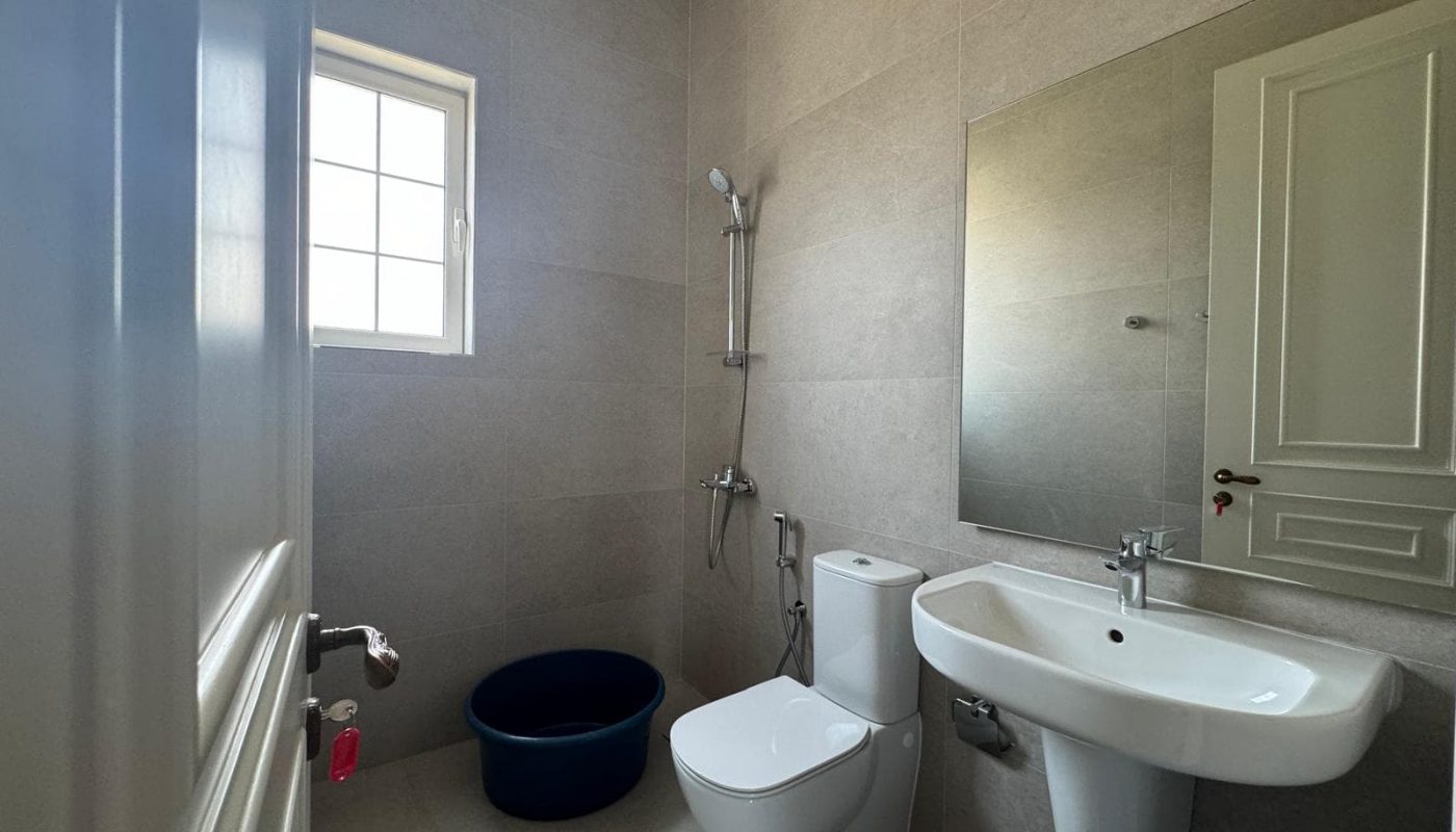 Bathroom with a sink, toilet, mirror, and wall-mounted showerhead. A small blue bucket is on the floor near the window. Walls are light grey tile.