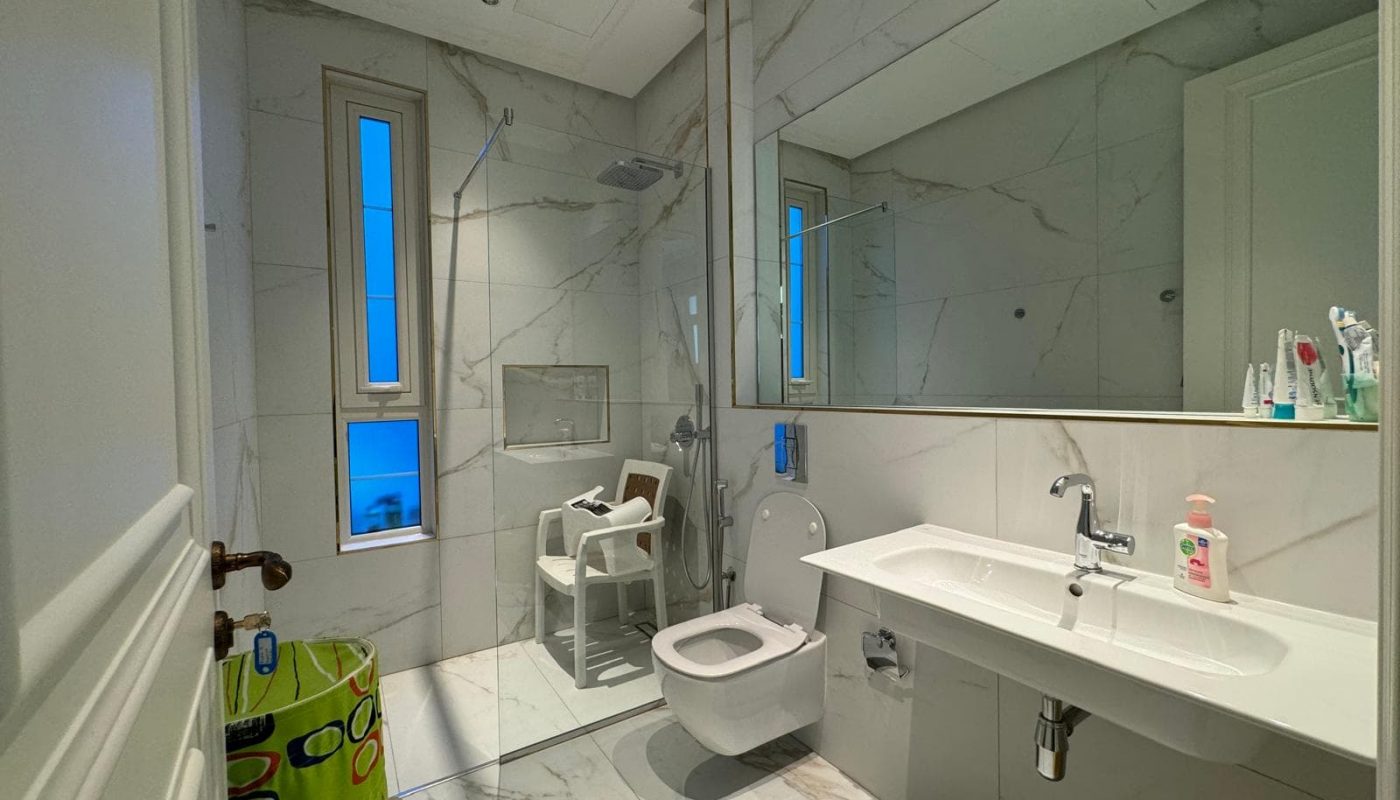 Modern bathroom with marble walls and floor. Features a glass shower enclosure, floating toilet, white sink, and a green wastebasket. Various toiletries are on the sink.