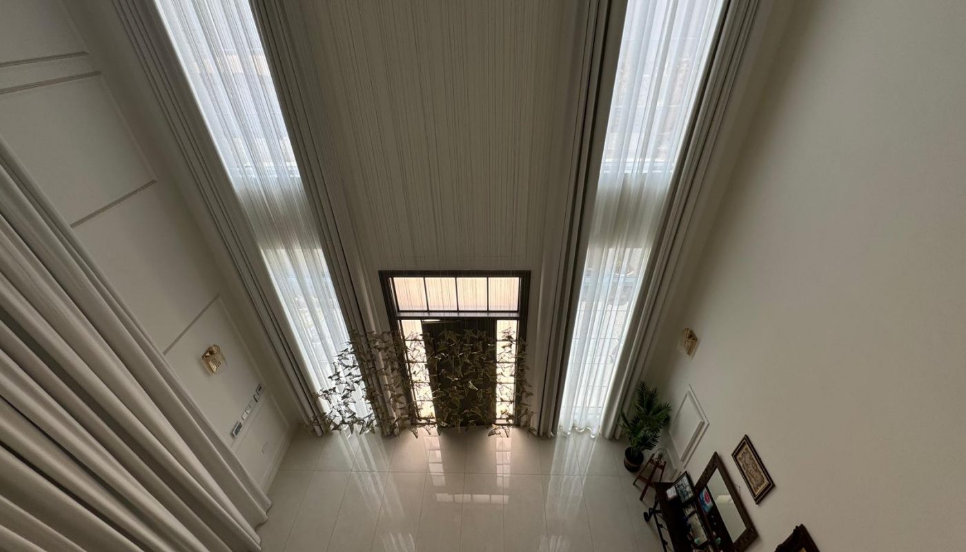 Interior view from a high angle showing tall windows with sheer curtains, a couch below, and a TV stand with framed pictures on the wall. The floor is tiled.