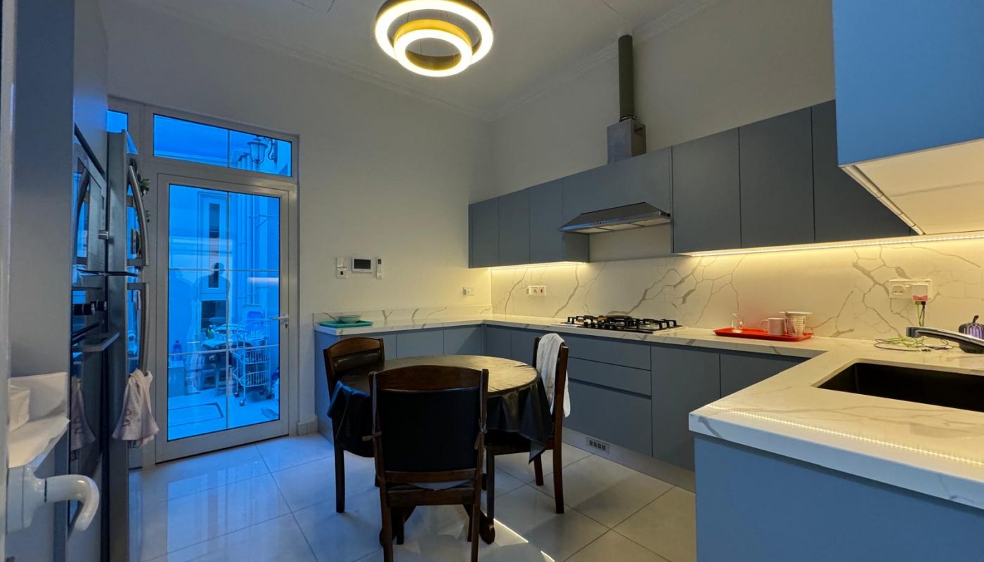 Modern kitchen with blue cabinets, built-in oven, and a round dining table with four chairs. A circular ceiling light is above the table. A small laundry area is visible through a glass door.