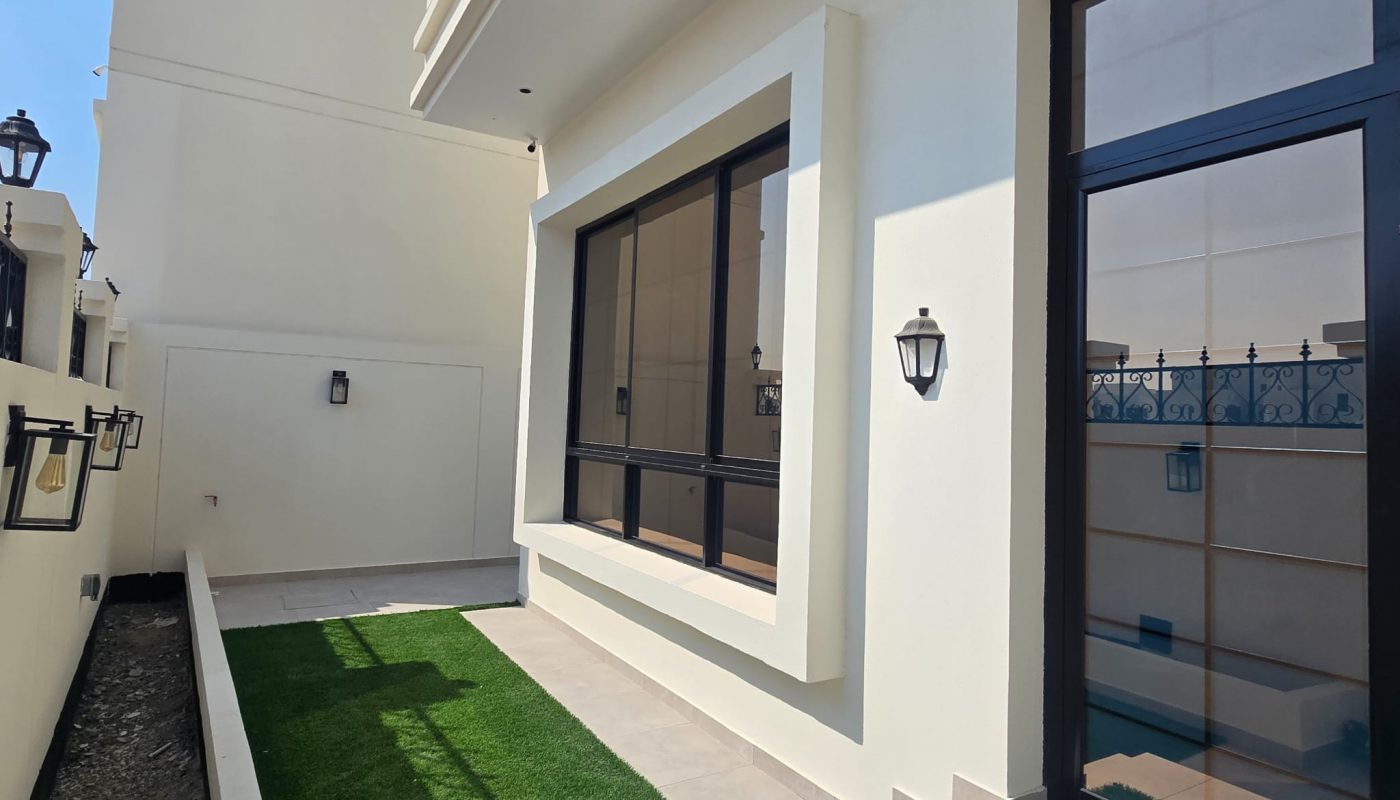 A modern house exterior with large windows, beige walls, wall-mounted lights, a grassy strip, and tiled steps under clear blue sky.