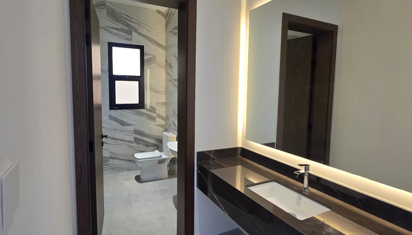 A modern bathroom with a long dark countertop and sink, large mirror with backlighting, and a view of a toilet and shower area through an open doorway.