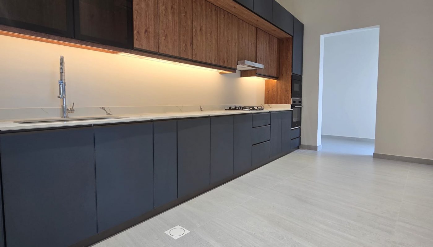 Modern kitchen with dark cabinets, wood accents, under-cabinet lighting, built-in oven, and light gray tile flooring. A doorway leads to an adjacent bright room.