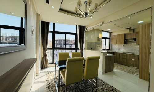 Modern dining area with a glass table and yellow chairs, adjacent to a kitchen with wooden cabinets and a chandelier, set against large windows with city views.