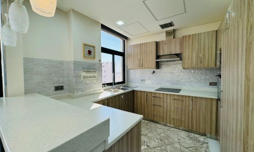 Modern kitchen with wood cabinets, light countertops, pendant lighting, and tiled flooring. White walls and a large window provide natural light.