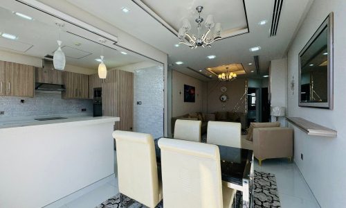 Modern open-plan dining and living area with a glass table and cream chairs, white kitchen, beige sofas, and a decorative chandelier on the ceiling.