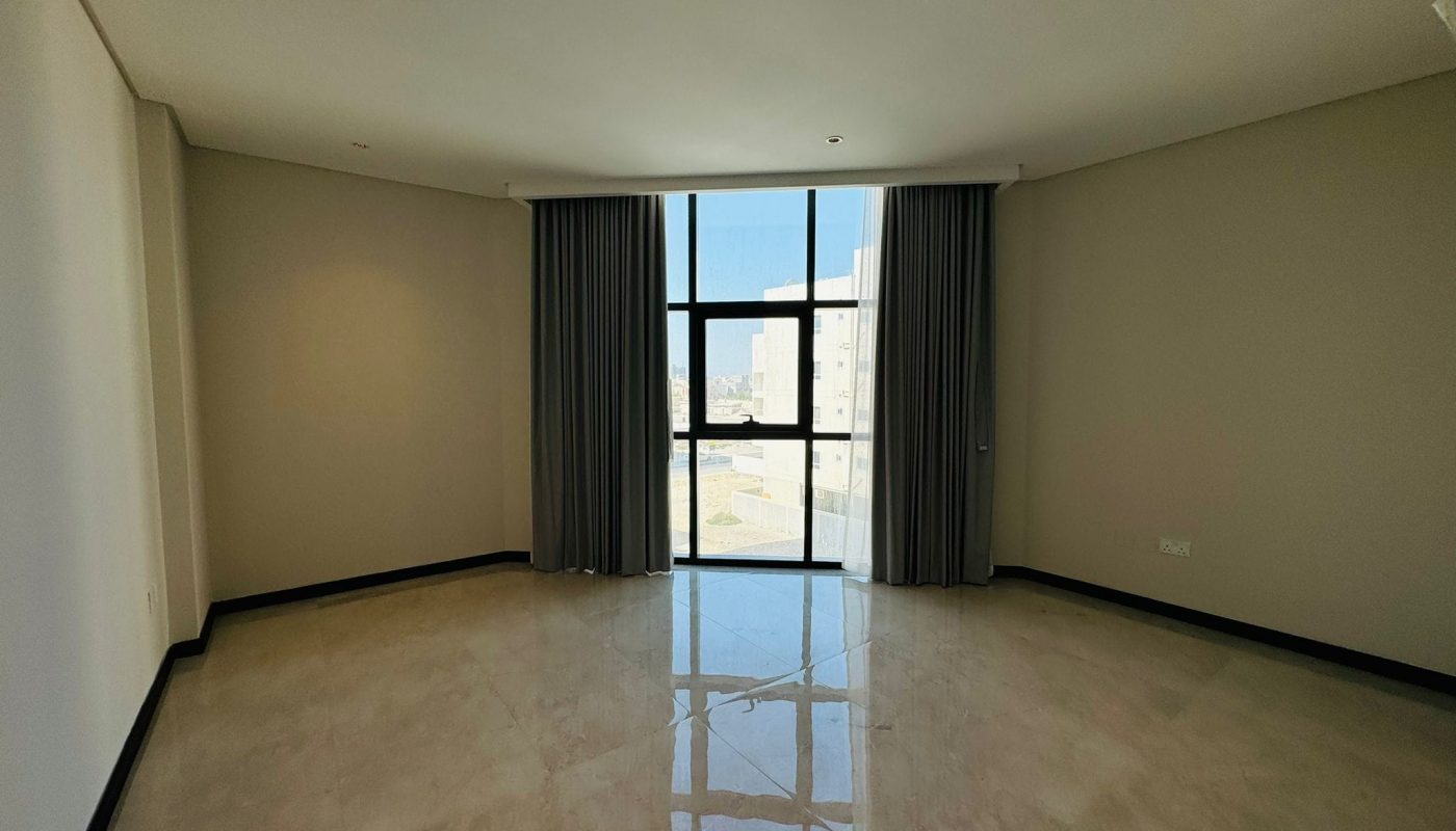 Empty room with a large window, gray curtains, and beige tiled floor. Sunlight reflects on the floor. Walls are painted light beige.