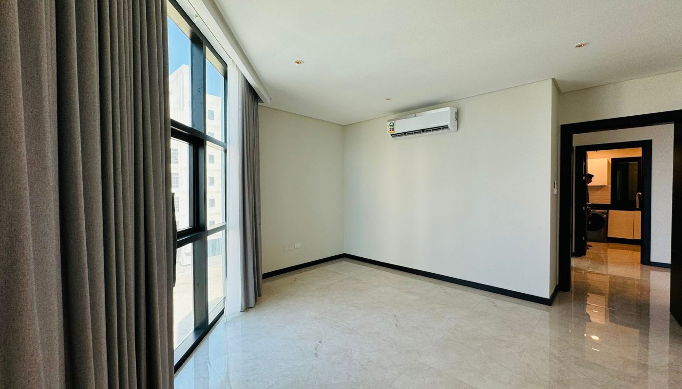 A bright, empty room with large windows, light-colored marble floors, gray curtains, and a wall-mounted air conditioning unit. A doorway leads to another area.