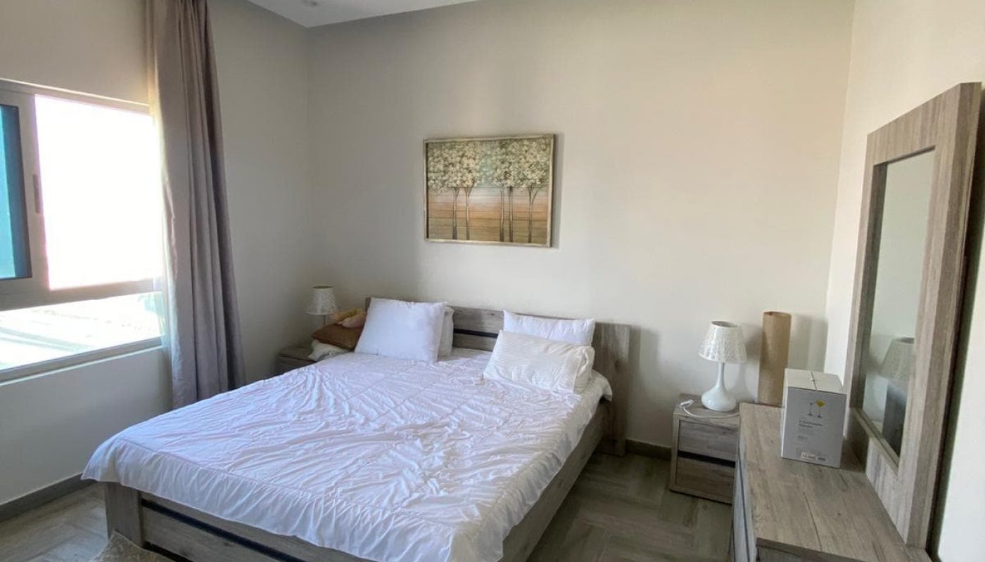 Bedroom with a neatly made double bed, two pillows, wooden furniture, a window with curtains, a wall picture, and a ceiling light fixture.