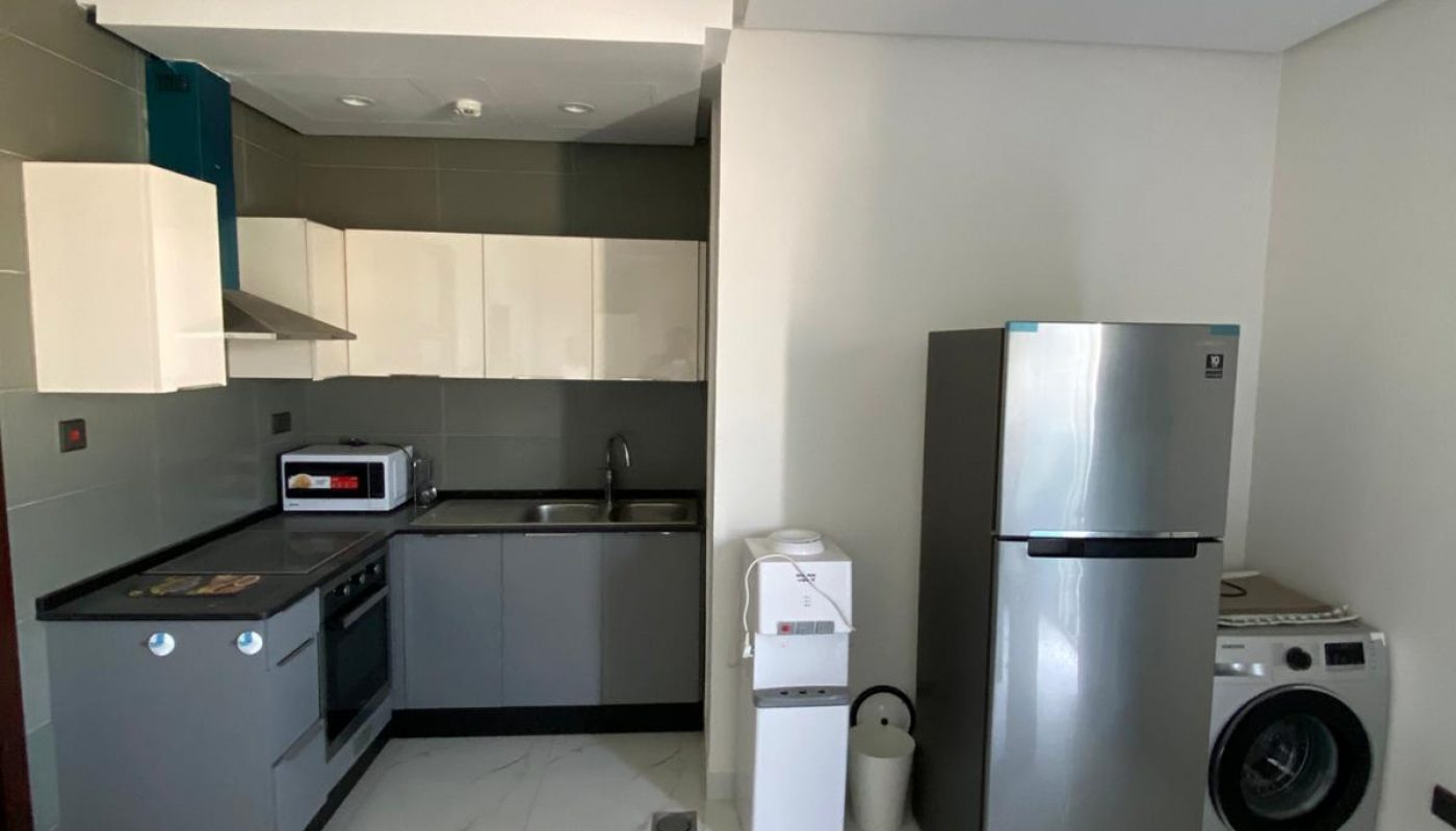 Modern kitchen with grey cabinets, stainless steel appliances, fridge, washing machine, microwave, and table with chairs. Light-colored flooring and walls create a bright atmosphere.