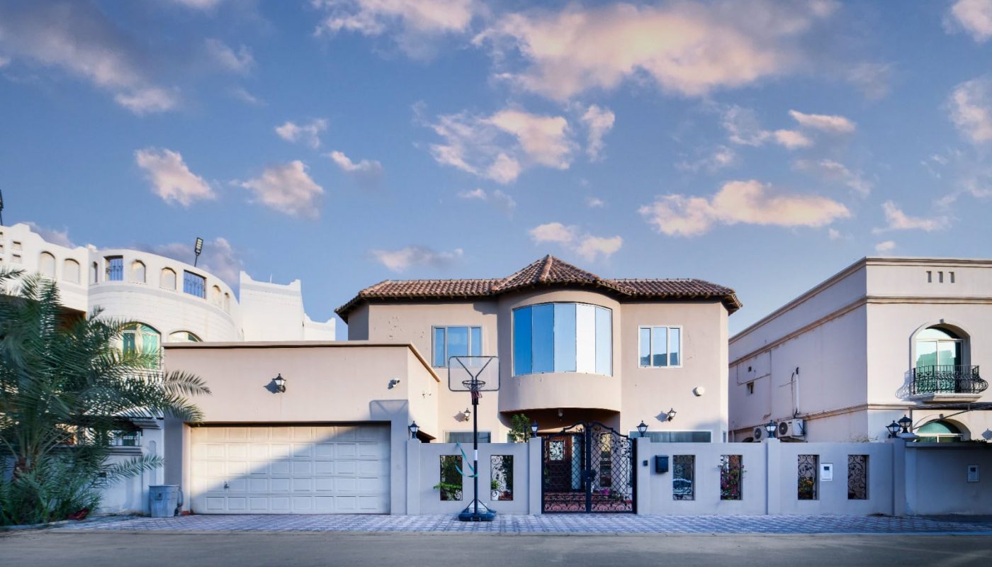 A two-story house with a tiled roof and a basketball hoop in the driveway. It's surrounded by a white fence, with nearby buildings. The sky is partly cloudy.