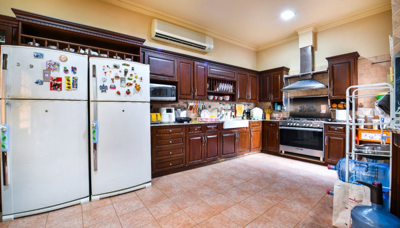A spacious kitchen with dark wooden cabinets, two refrigerators with magnets, a large stove, microwave, and various kitchen items. The floor is tiled, and there are water dispensers on the side.