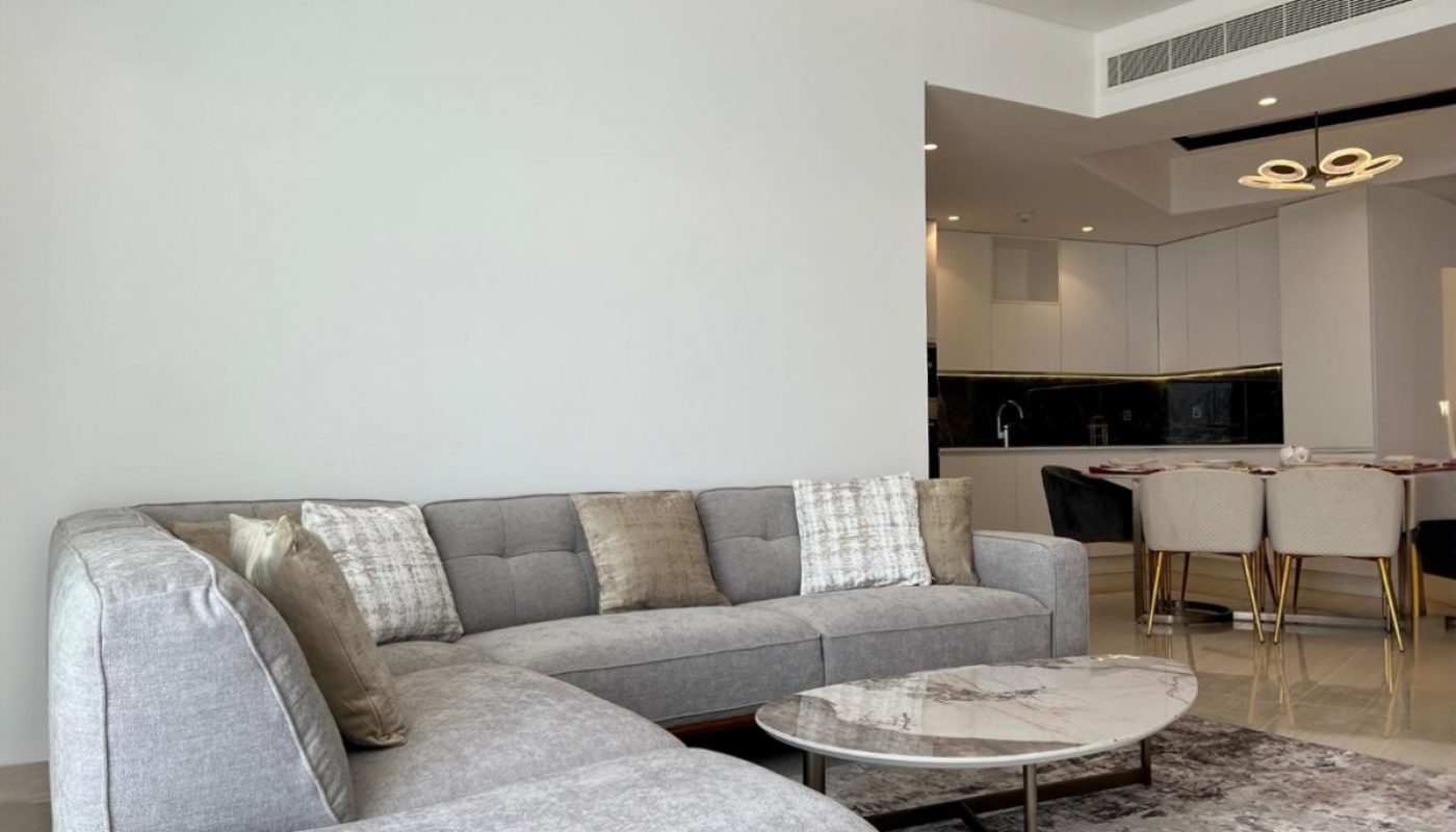 Modern living room with a gray sectional sofa, marble coffee table, neutral rug, and open kitchen layout with dining area in the background.