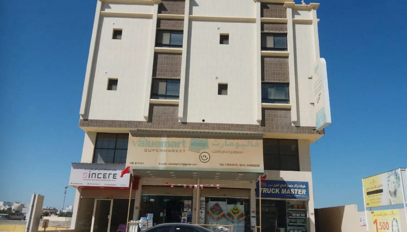 A three-story beige building with a supermarket, a shop, and a truck business on the ground floor; a gray car and orange cones are parked in front.