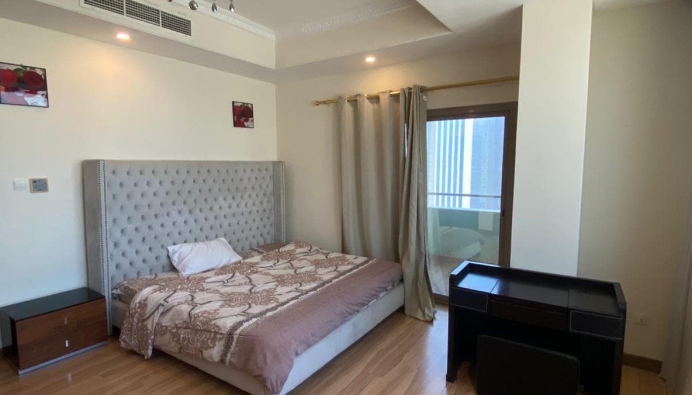 Bedroom with a large bed, brown bedding, two nightstands, and a desk. Curtains cover a window. Chandelier on the ceiling and two small paintings hang on the walls.