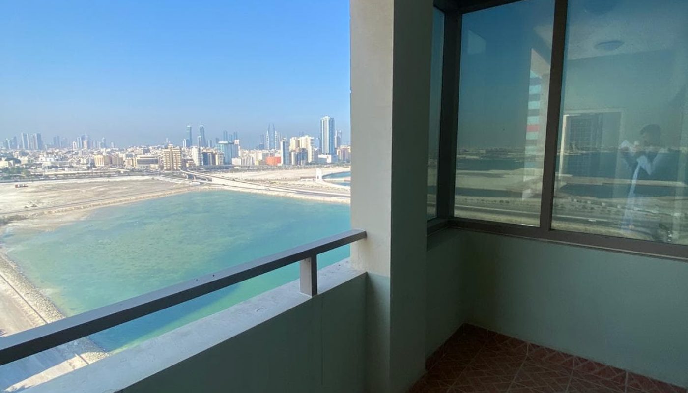 Balcony view of a cityscape with tall buildings, overlooking a body of water and shoreline. The balcony has tiled flooring and is partially enclosed with a railing and walls.
