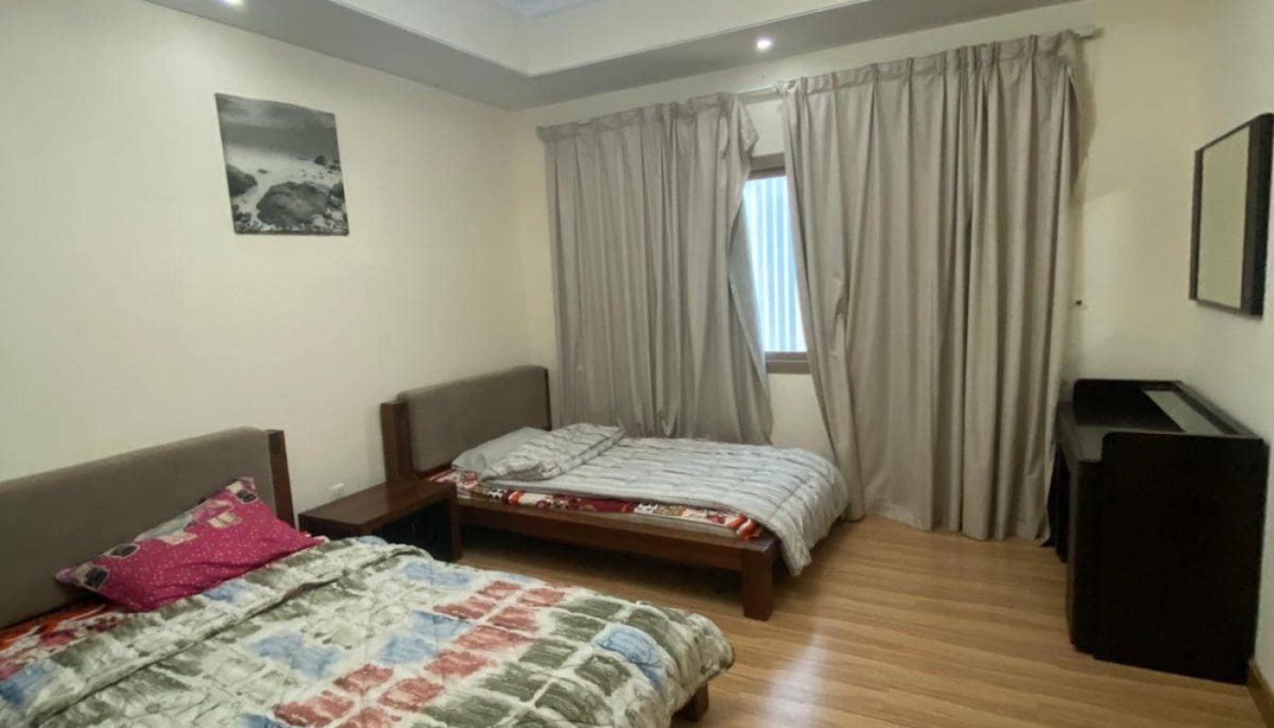 A bedroom with two beds, each with patterned bedding. The room has wooden flooring, a window with curtains, a ceiling light, and a wall mirror.