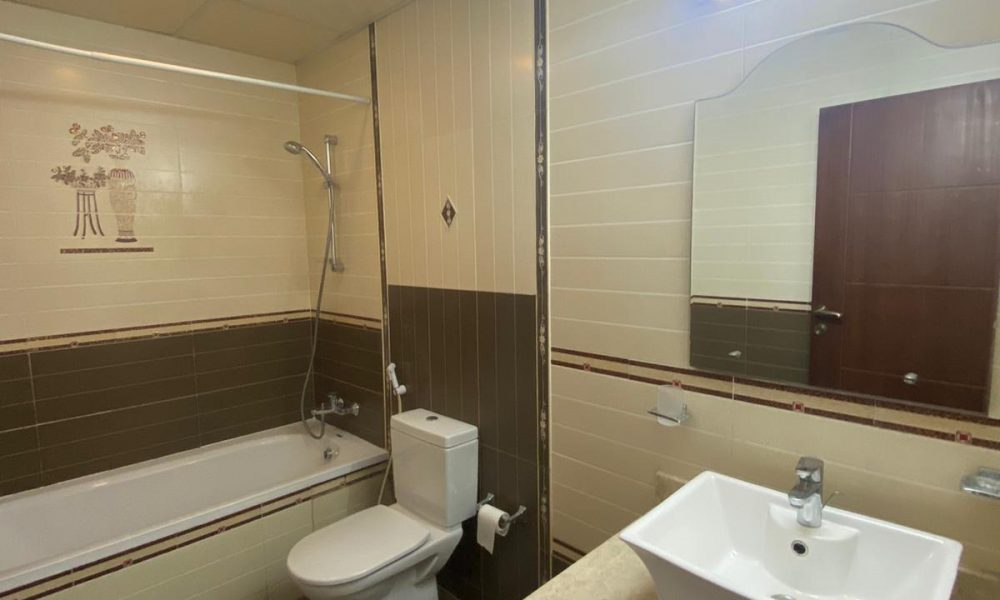 A bathroom with a bathtub, shower, toilet, and sink. There's a wall-mounted mirror above the sink and a light fixture above it. The walls and floor have light and dark tile patterns.