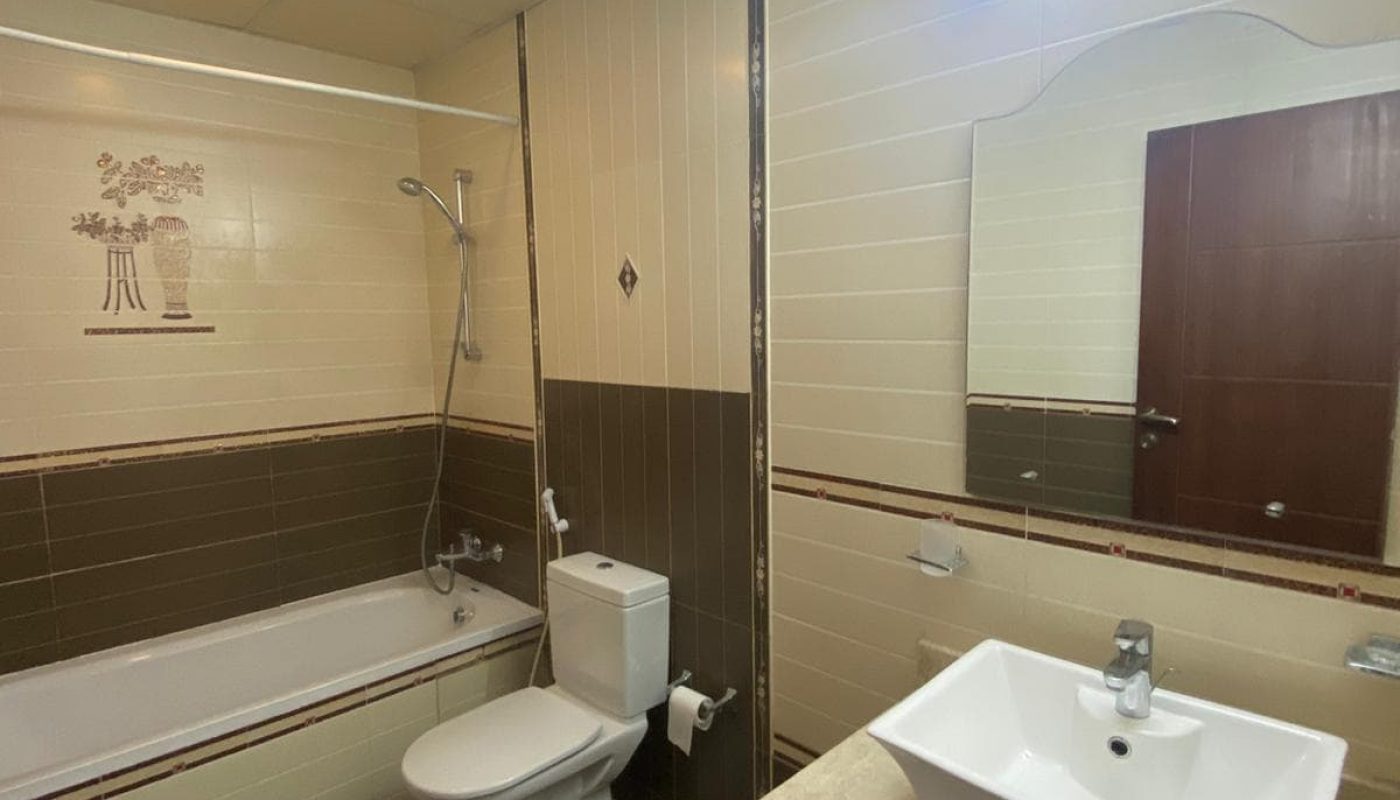 A bathroom with a bathtub, shower, toilet, and sink. There's a wall-mounted mirror above the sink and a light fixture above it. The walls and floor have light and dark tile patterns.