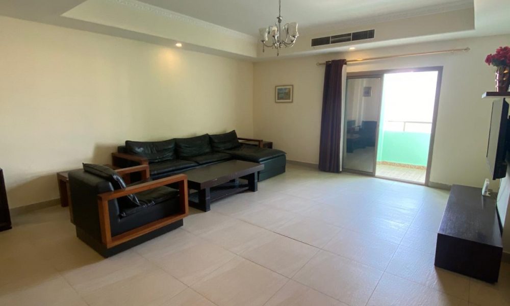 Spacious living room with a black leather sofa set, coffee table, and a TV on a stand. Large window with curtains and a small balcony visible in the background.