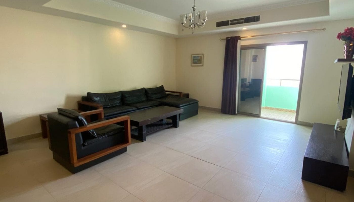 Spacious living room with a black leather sofa set, coffee table, and a TV on a stand. Large window with curtains and a small balcony visible in the background.