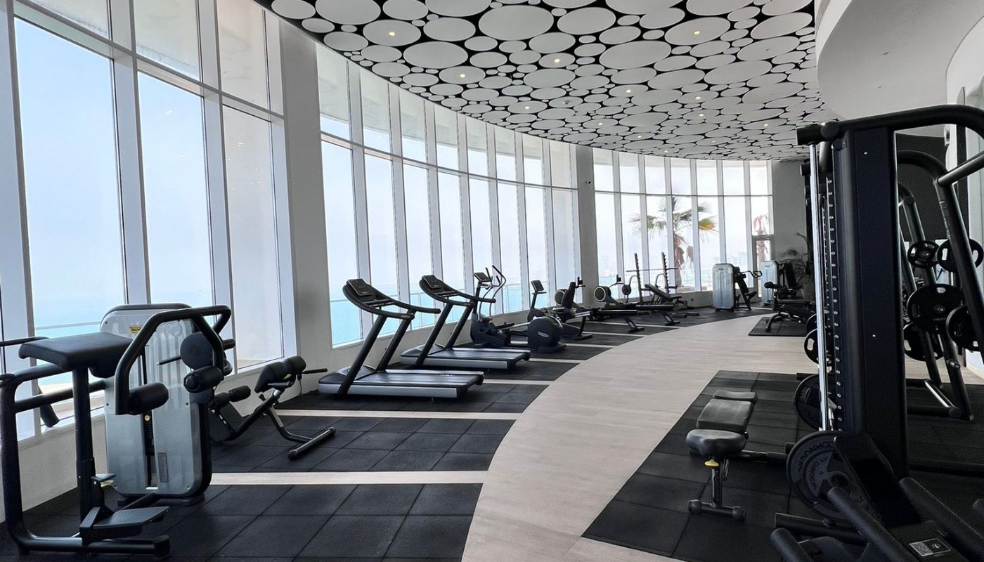 Modern gym with treadmills, rowing machine, and weight equipment, featuring large windows and a ceiling with circular patterns.