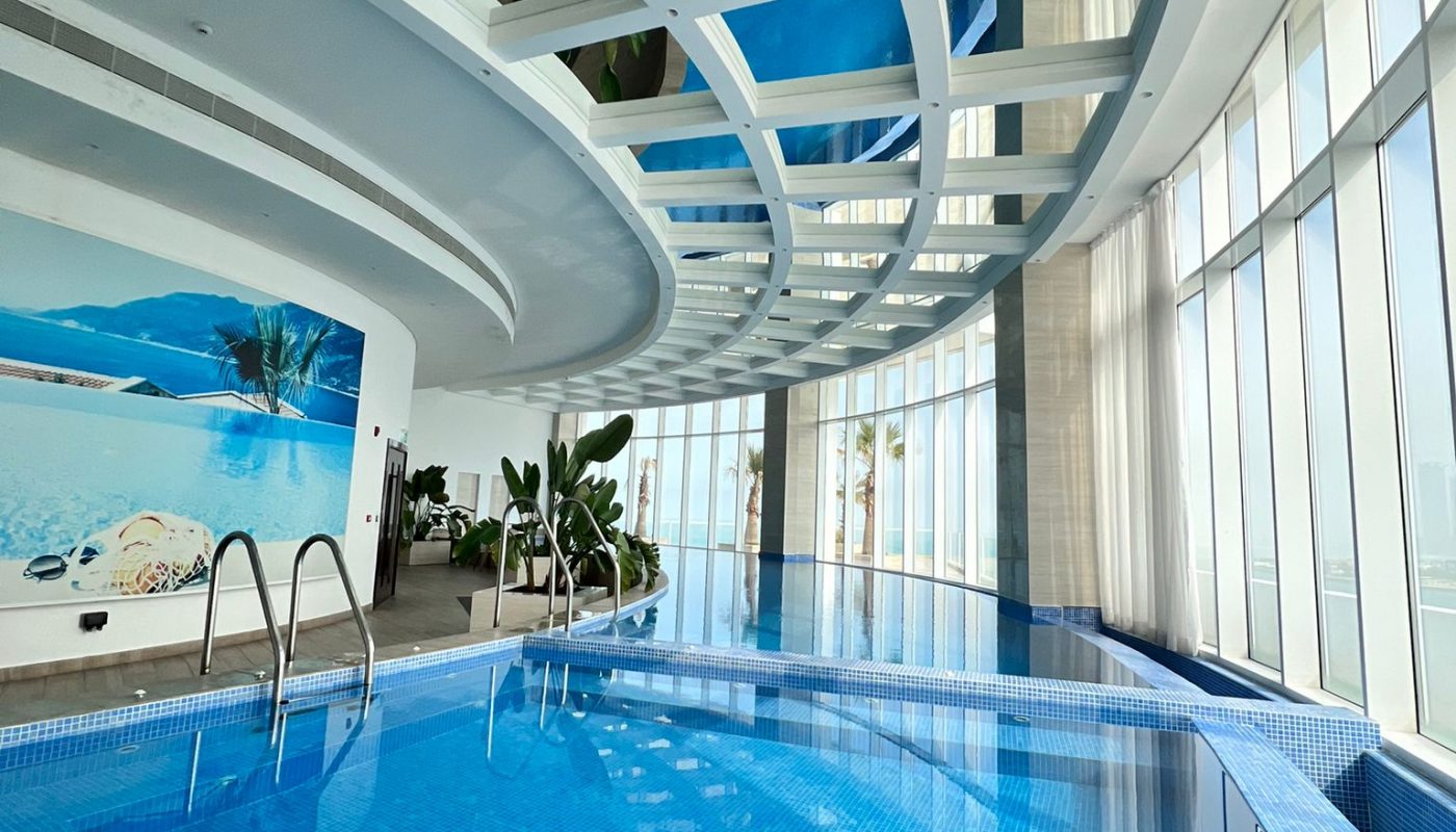 Indoor swimming pool with blue tiles, large curved glass windows, a sloped ceiling with skylights, and a wall mural of a beach scene. Steps and plants are visible near the pool.