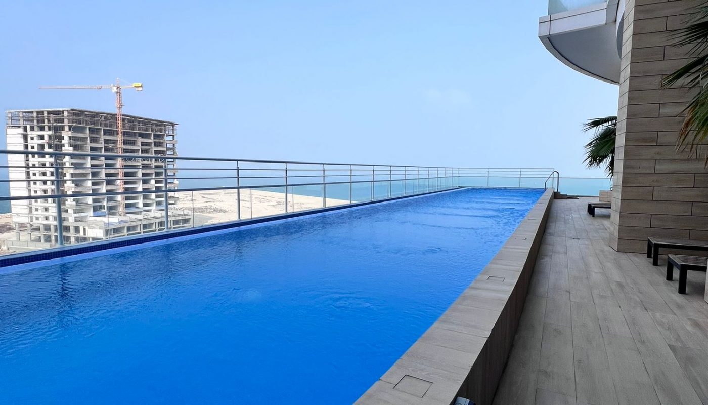 A long rooftop swimming pool with a view of the ocean and a nearby construction site, adjacent to a modern building with balconies.