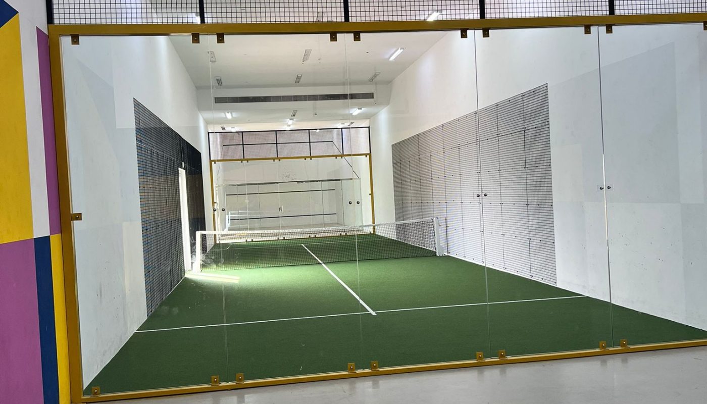 Indoor sports court with artificial turf, glass walls, and overhead netting; white walls with some areas painted in bright colors on the left.