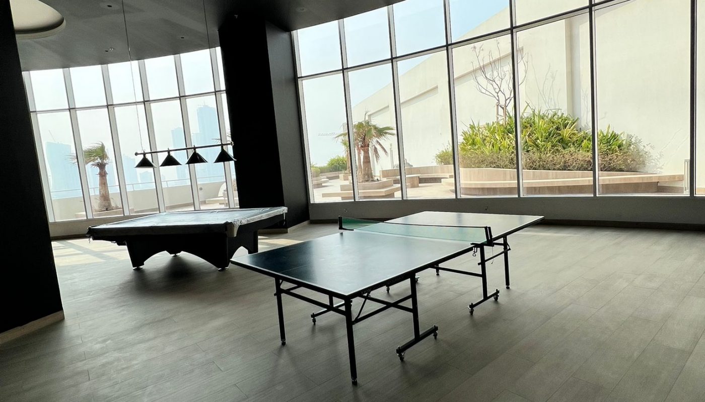 A room with large windows features a pool table and a ping pong table, with natural light coming in and a view of plants outside.