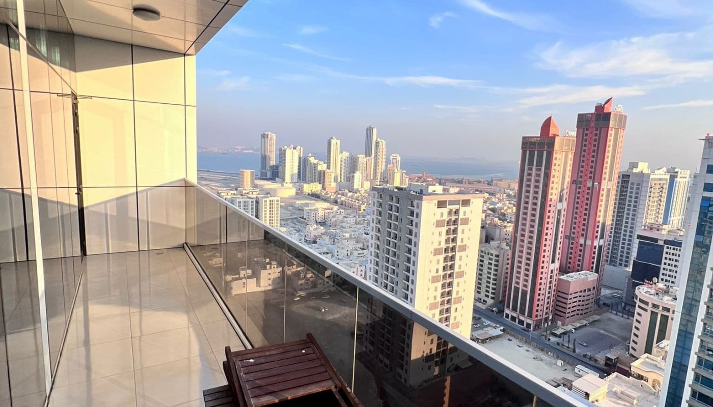 Balcony view from a high-rise building, overlooking a cityscape with tall buildings under a blue sky. A wooden chair sits on the shiny tiled floor of the balcony.