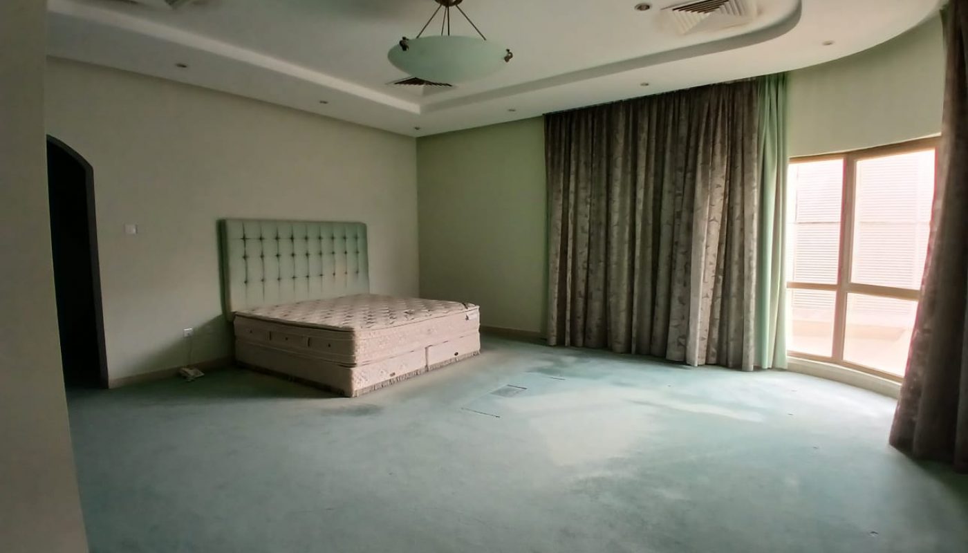 Large, empty bedroom with a green carpet, a bed without bedding, curtains covering windows, and a modern ceiling light fixture.