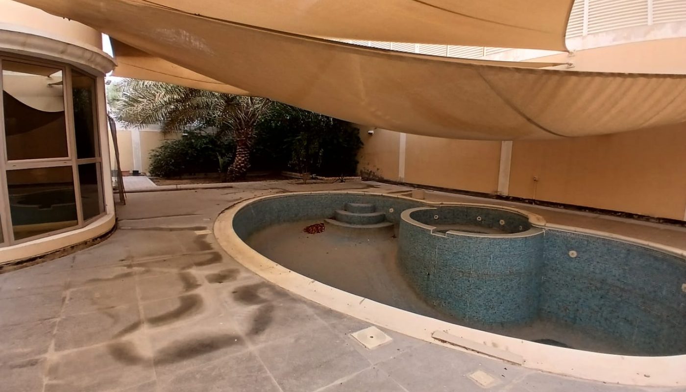 An empty, tiled outdoor pool and jacuzzi under large beige shade sails, with dirt on the floor and some plants in the background.