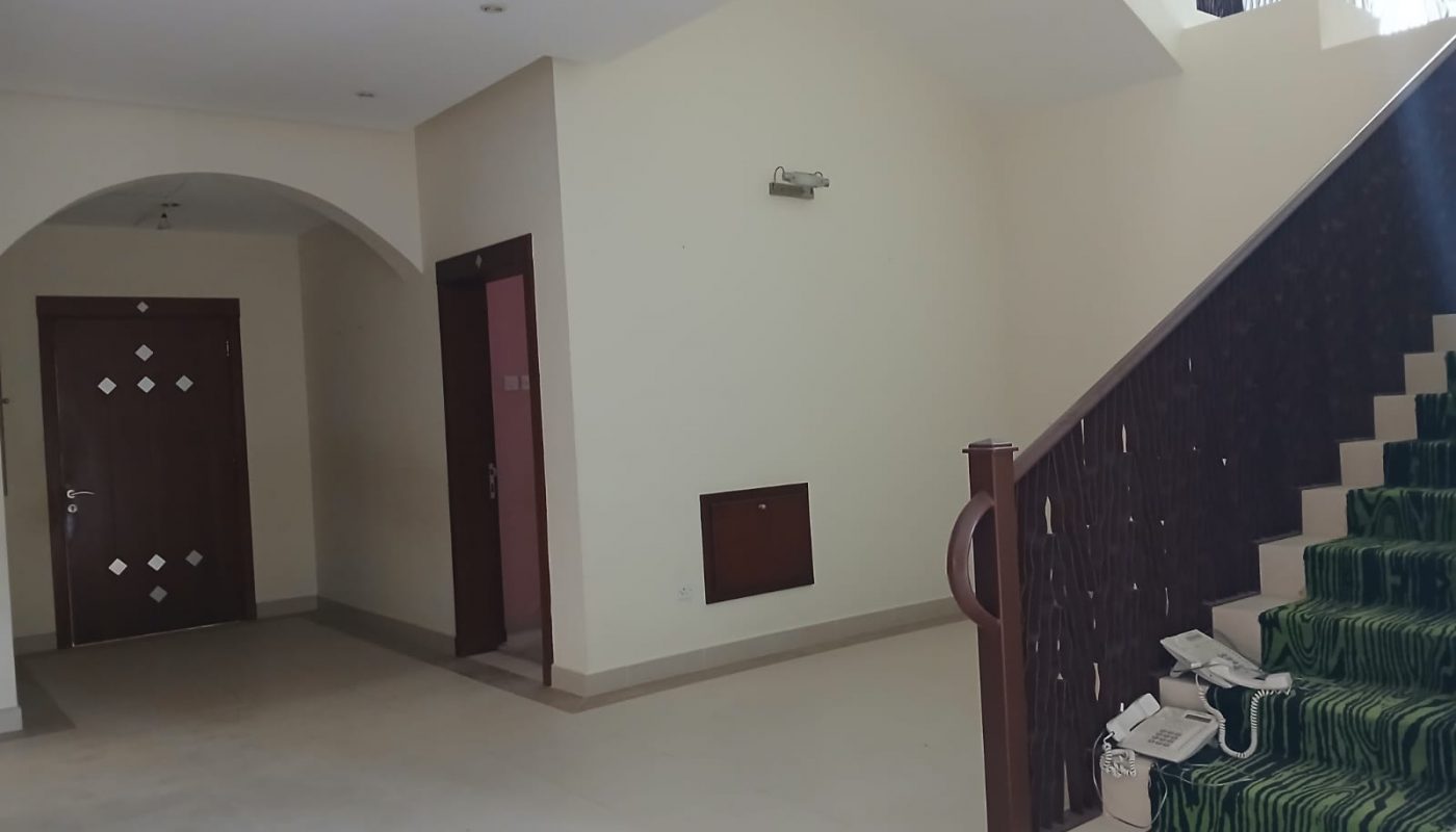 A staircase with green carpet and wooden railing, adjacent to beige walls and a brown door in a spacious, unfurnished interior hallway.