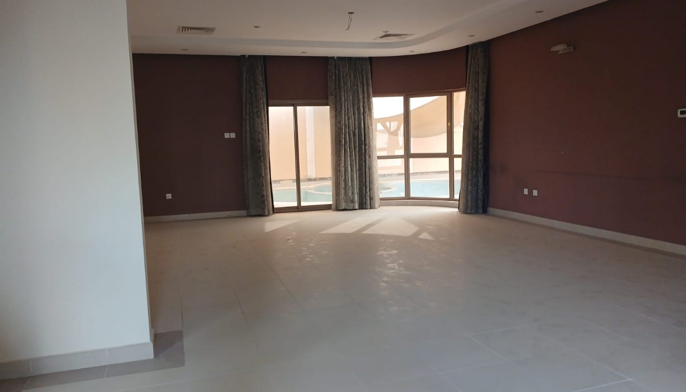 Spacious empty room with tiled floor, brown accent wall, large windows and glass door view of a pool outside, and long curtains.
