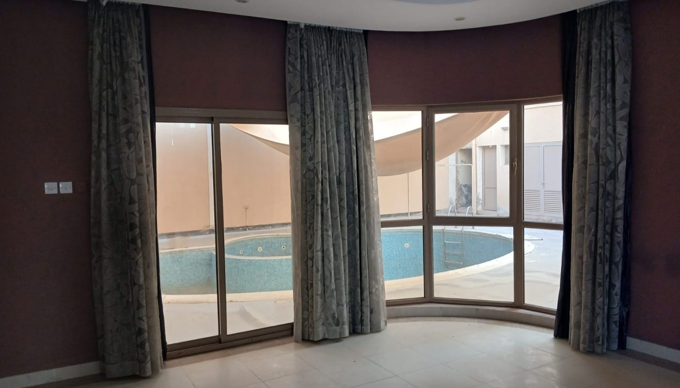A room with tiled floor, large windows, and heavy curtains overlooks a small outdoor swimming pool surrounded by beige walls.