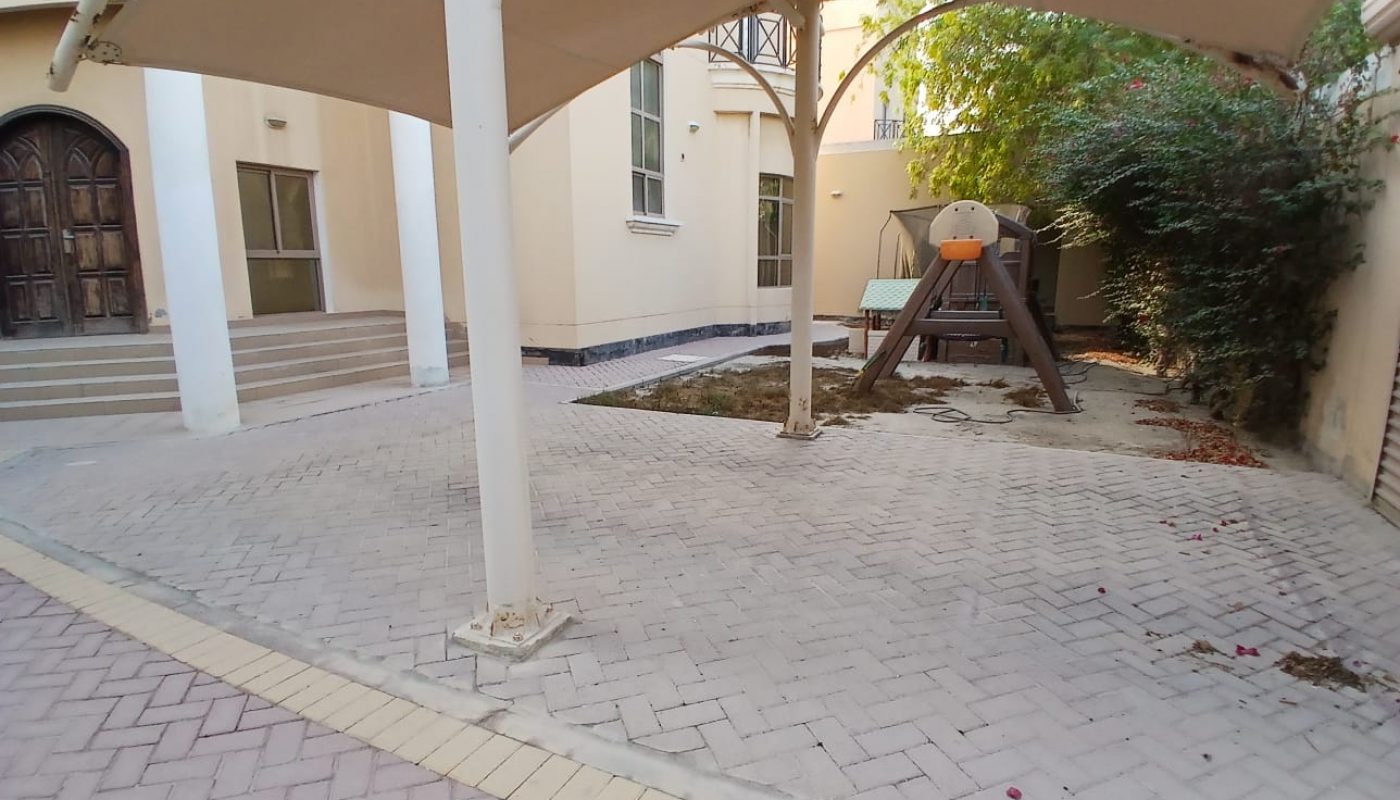 A paved courtyard with a beige canopy shade, a wooden play structure with a slide, some greenery, and a building with arched doors and windows in the background.
